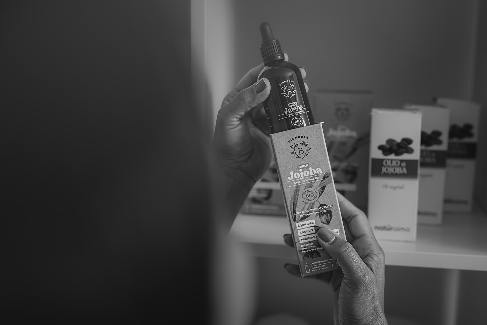 Hand houdt een fles jojoba-olie omhoog, met verpakkingen op de achtergrond in een grijszwarte sfeer.