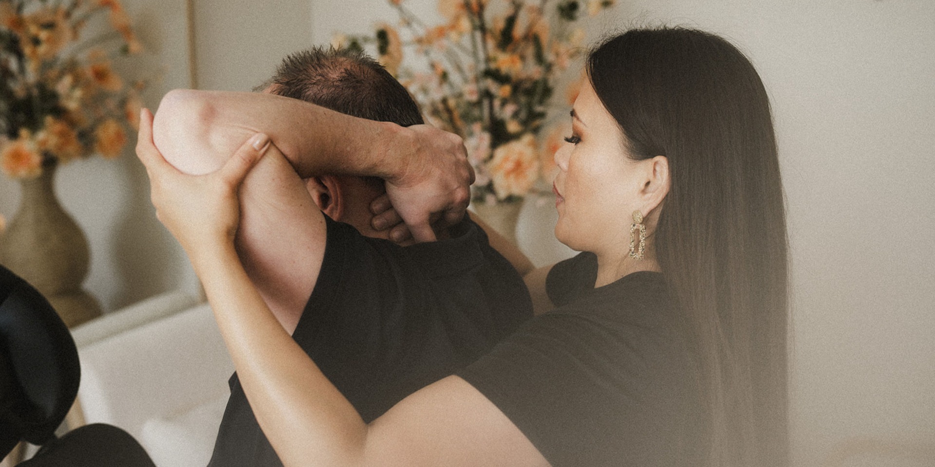 Man op stoelmassage krijgt massage van vrouw, in lichte kamer met bloemenvaas op achtergrond.